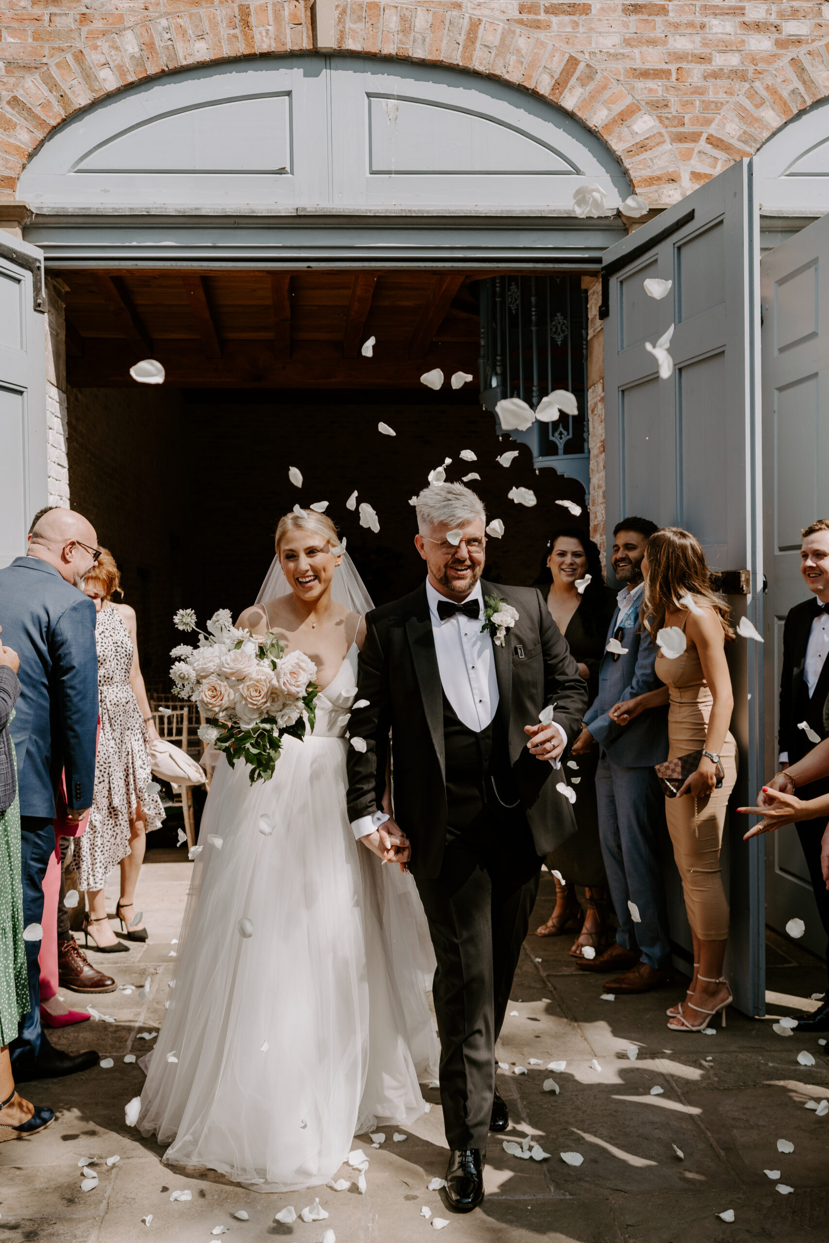 A bride, holding a bouquet, and groom exiting a wedding ceremony with confetti thrown at them, with a row of wedding guests either side.