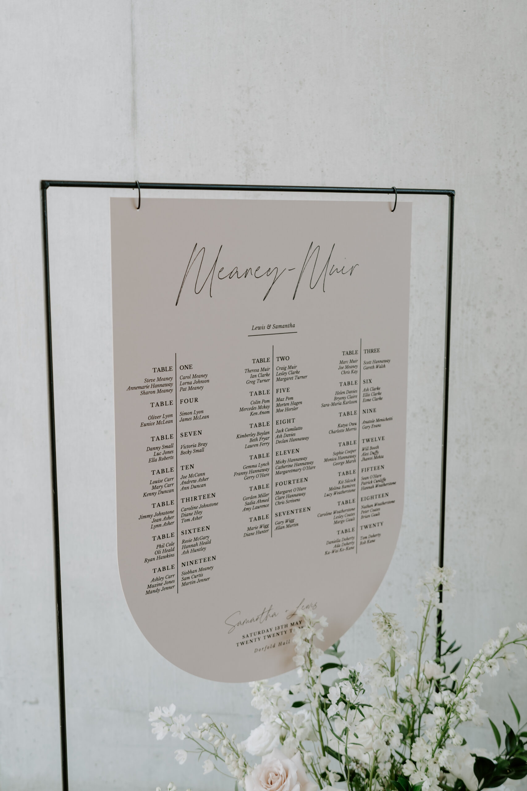 A wedding seating chart and table plan on a thin black wrought iron stand with flowers at the base.