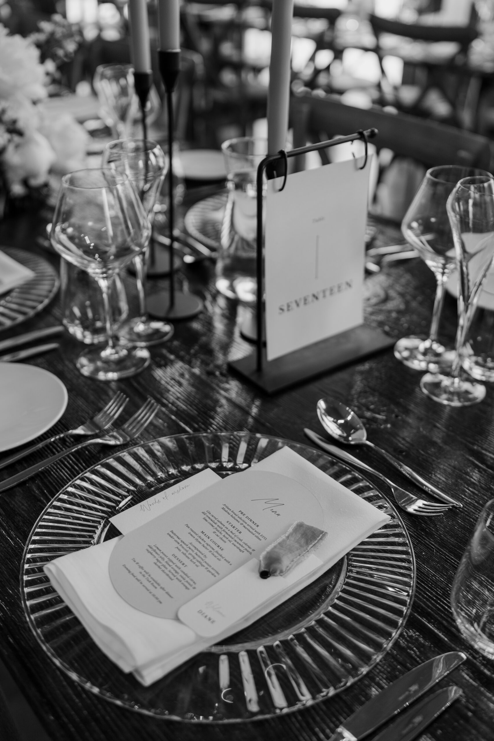 A black and white photo of a beautifully styled wedding table place setting with clear glass plate, wedding menu and silverware.