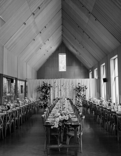 A black and white photo showcasing long wedding tables styled with place settings and flowers, set in a hall.