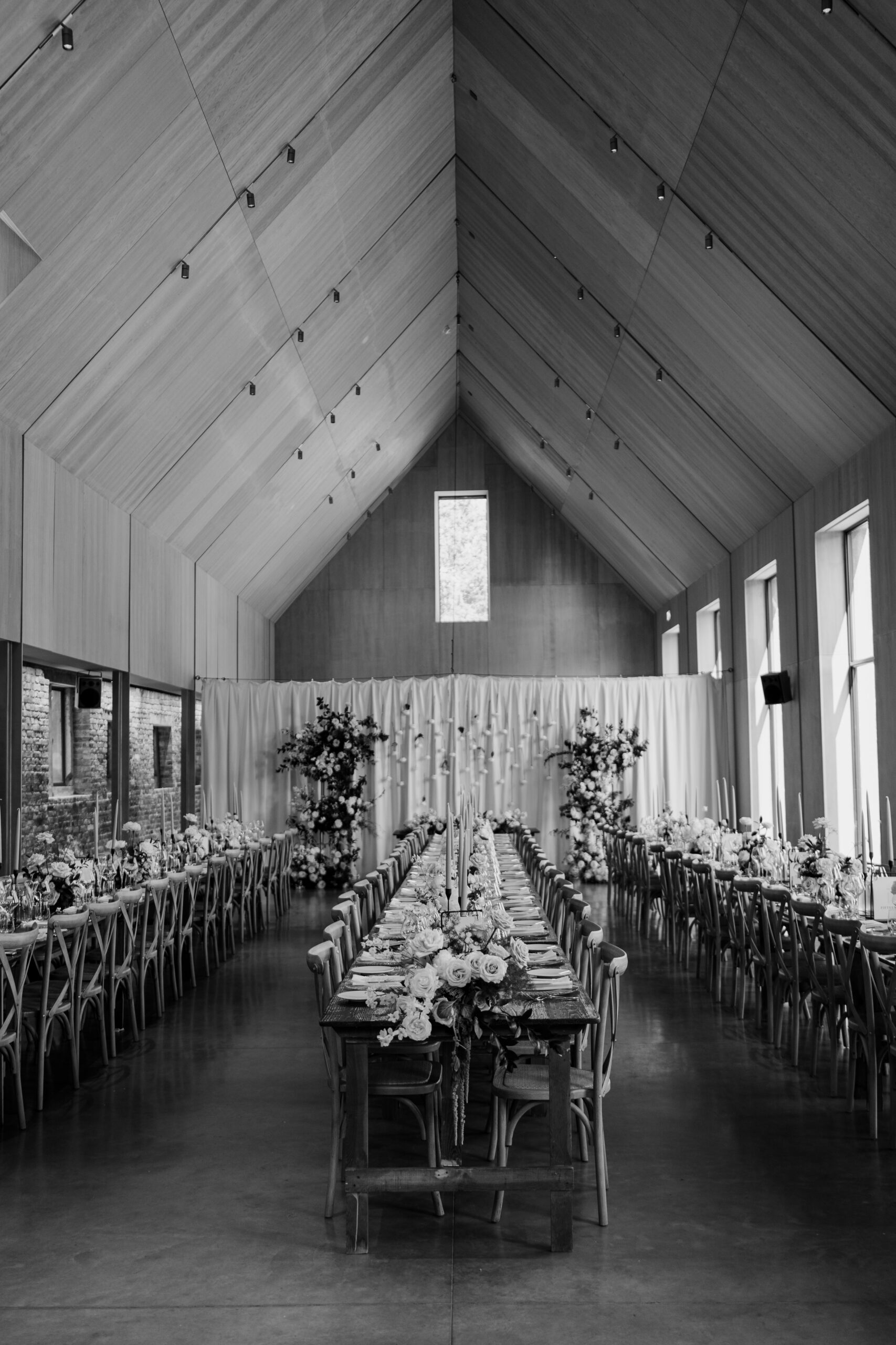 A black and white photo showcasing long wedding tables styled with place settings and flowers, set in a hall.