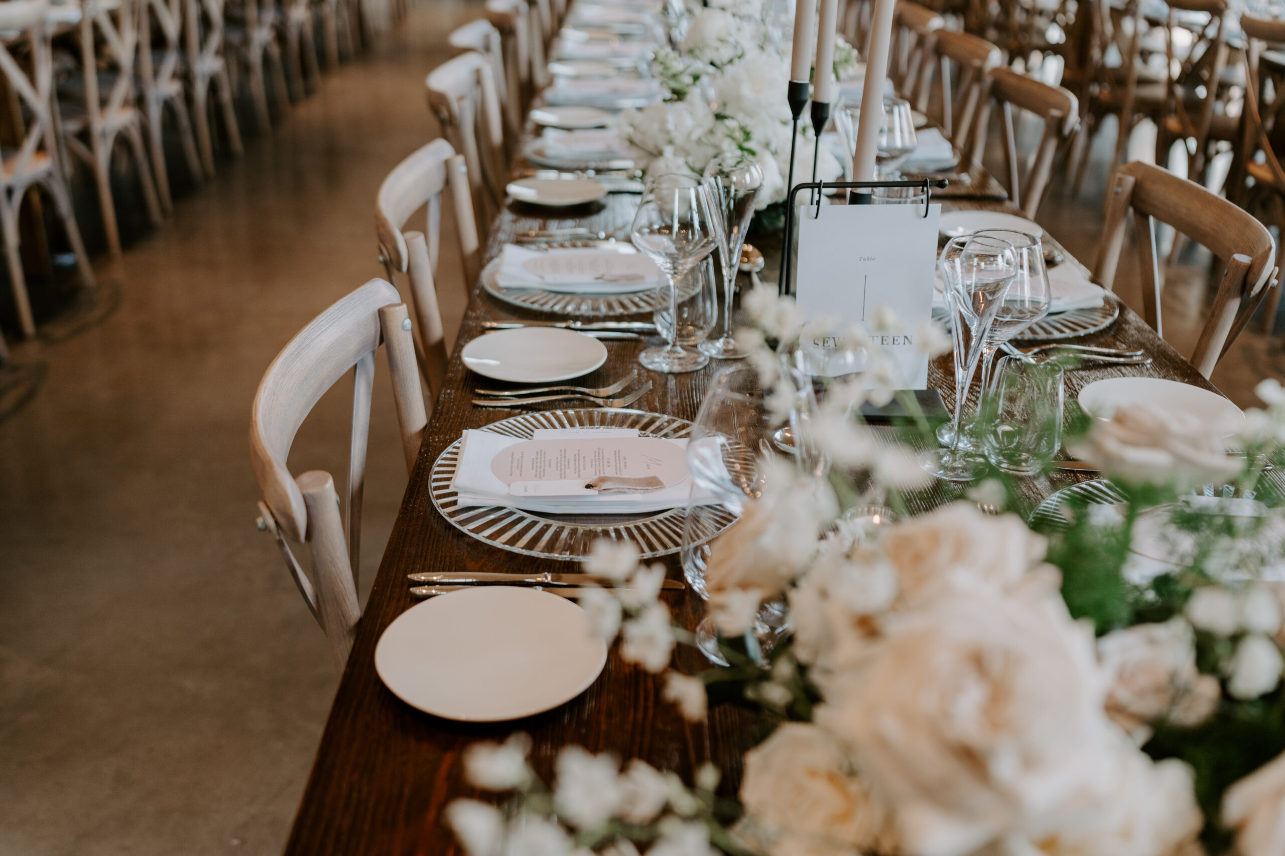 A beautifully styled long table with white flowers, candles and place settings and a table number card.