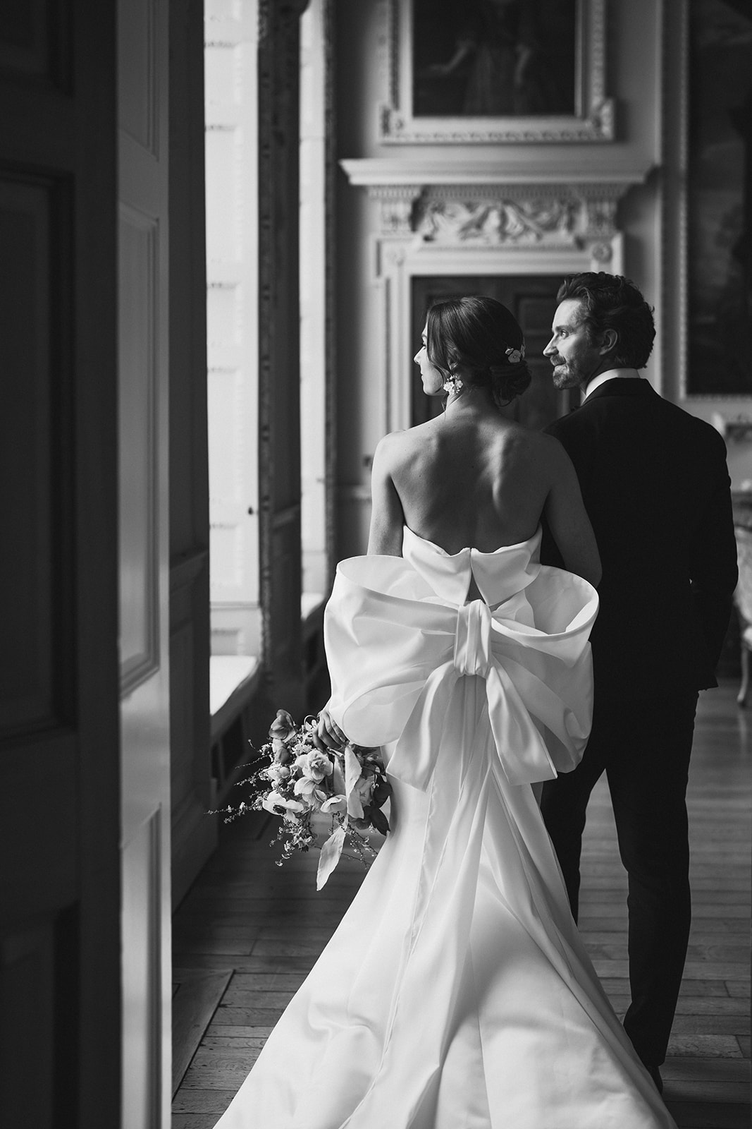 Hawkstone Hall Editorial // Petite Dauphine A black and white image of a bride with her back facing us with a large bow on the back of her wedding dress. She is looking out of one of the large windows at Hawkstone after her wedding ceremony.