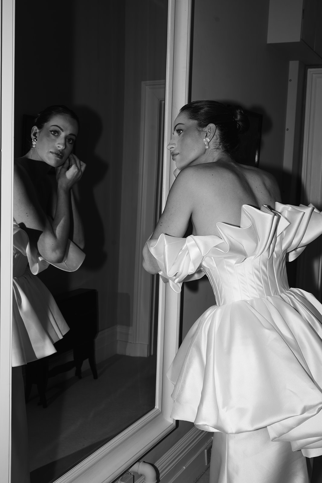 Birdsall House A black and white image of a bride putting her earrings on in front of a mirror and looking directly at the camera at Birdsall House in Yorkshire.