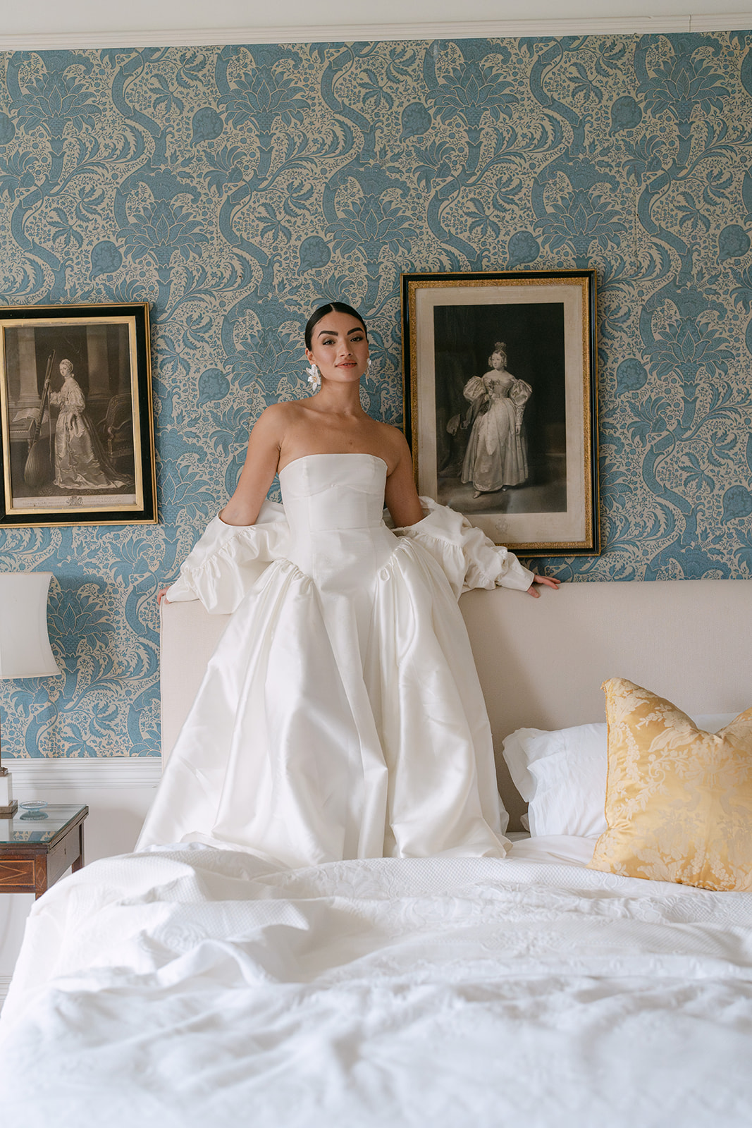 Birdsall House A colour image of a bride standing on a bead in her her wedding dress with original William Morris wallpaper behind her in a soft blue colour.