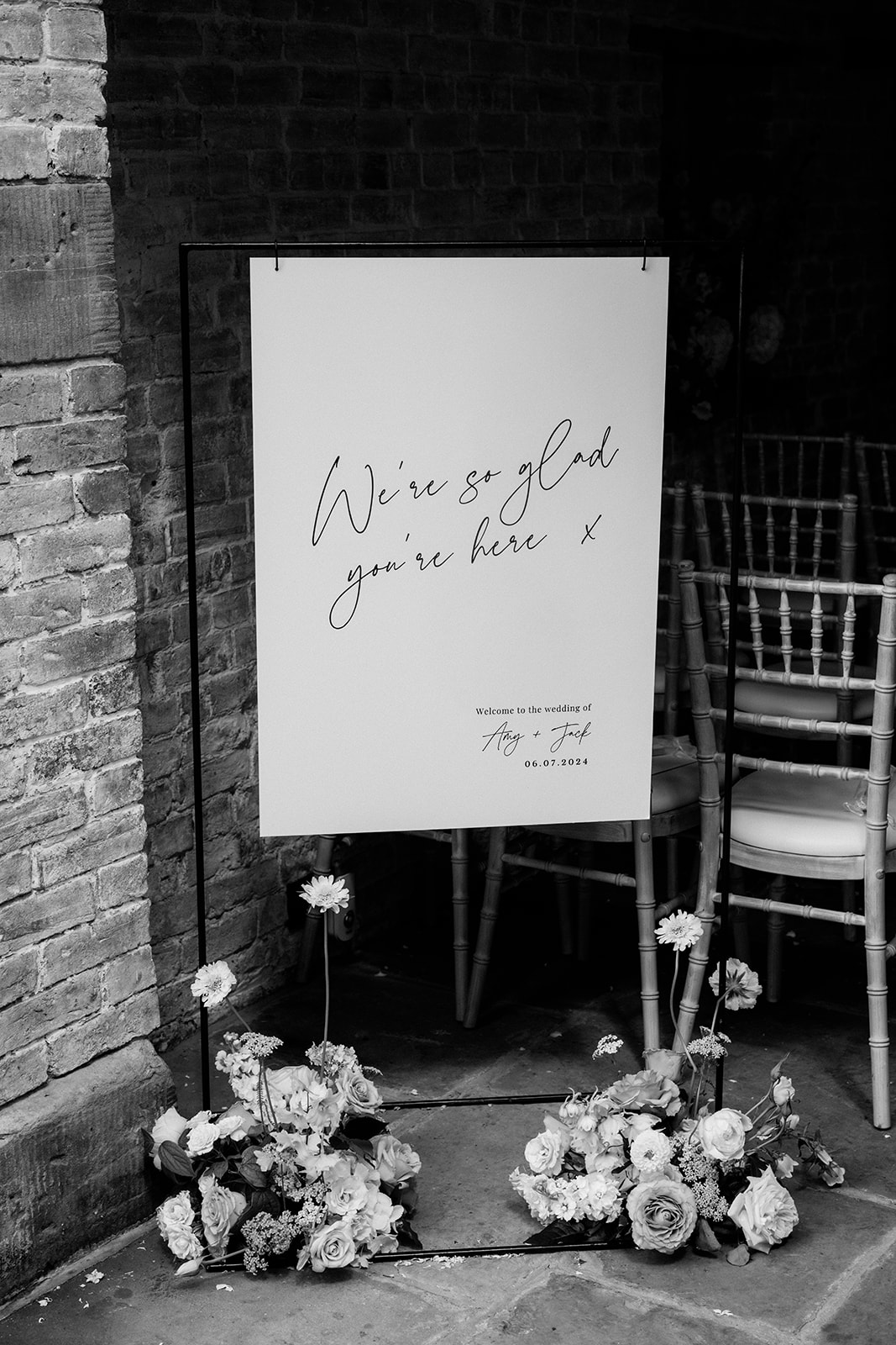 Amy and Jack A black and white image of a wedding welcome sign with flowers by sass flower at the base at a wedding ceremony at Dorfold Hall venue in Cheshire.