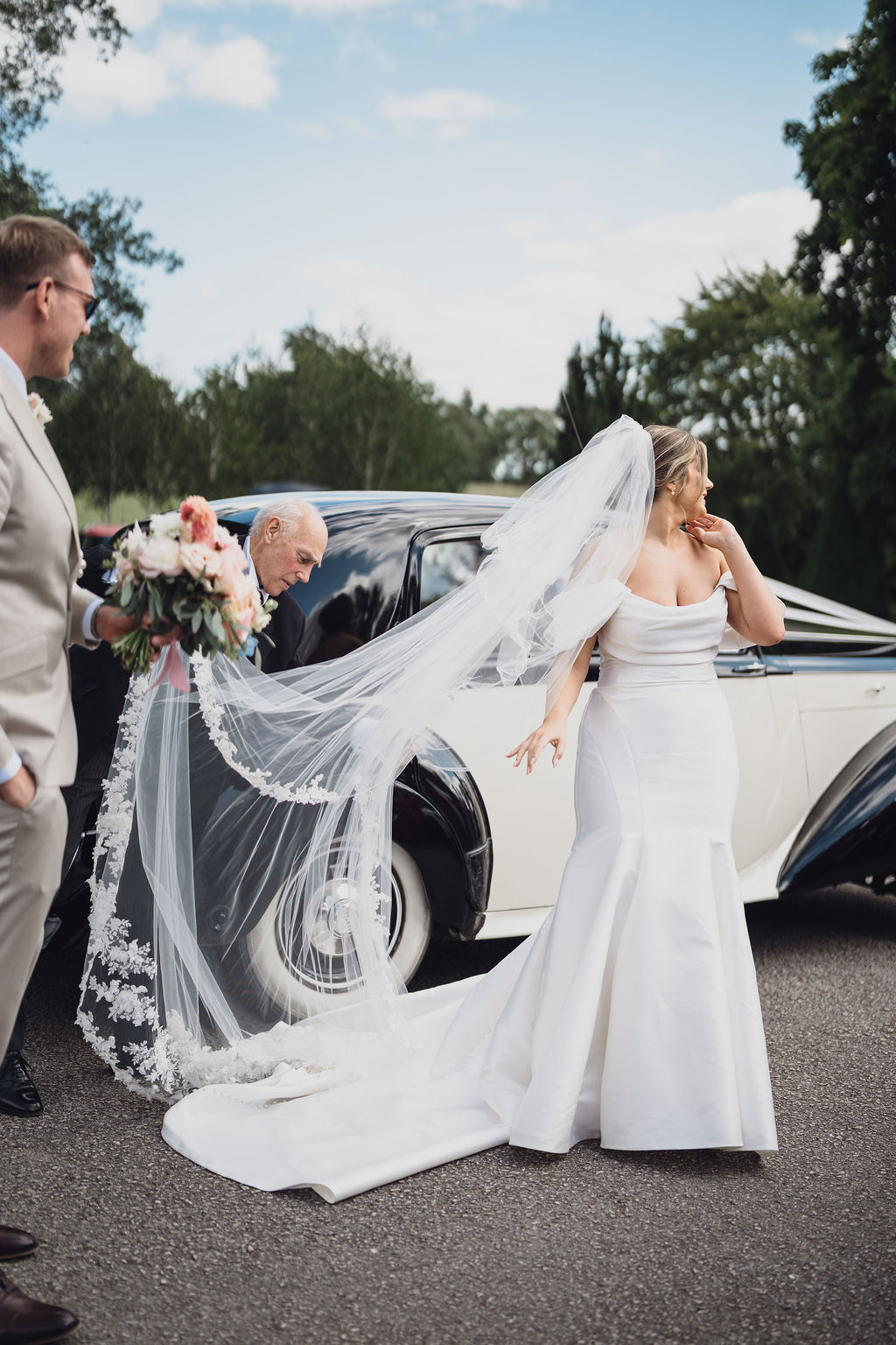 A colour image of a bride getting out of her wedding car with a beautiful lace veil.
