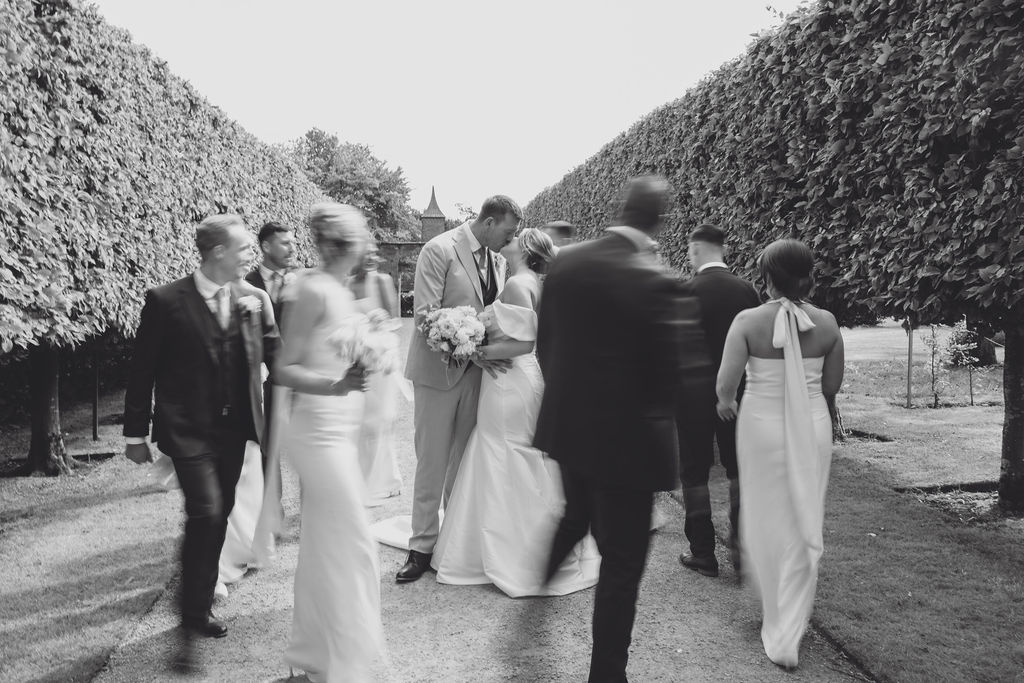 A black and white editorial style image of a bridal party circling the bride and groom as they kiss at Combermere Abbey Shropshire.