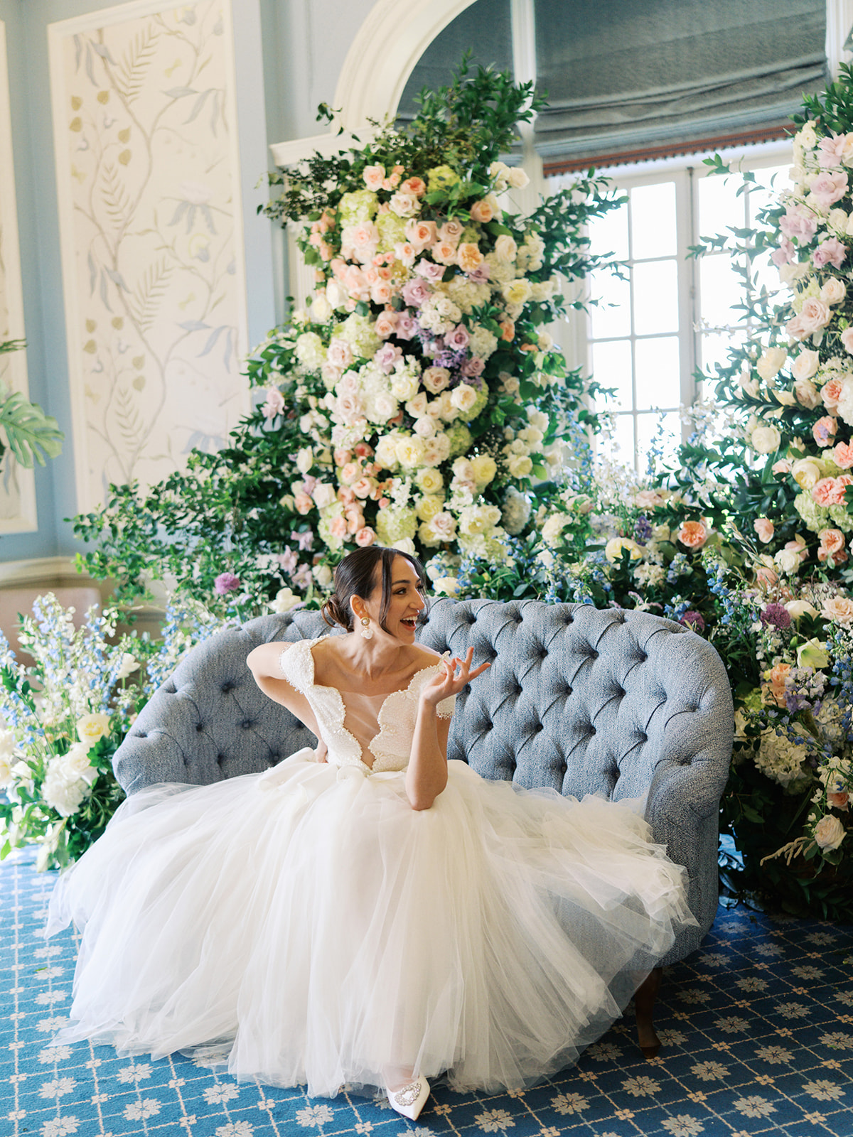 A colour image of a bride sat on a blue sofa in from of a huge spring floral installation at Kin House UK wedding venue. Wedding designed by Soir&eacute;e Studio. 